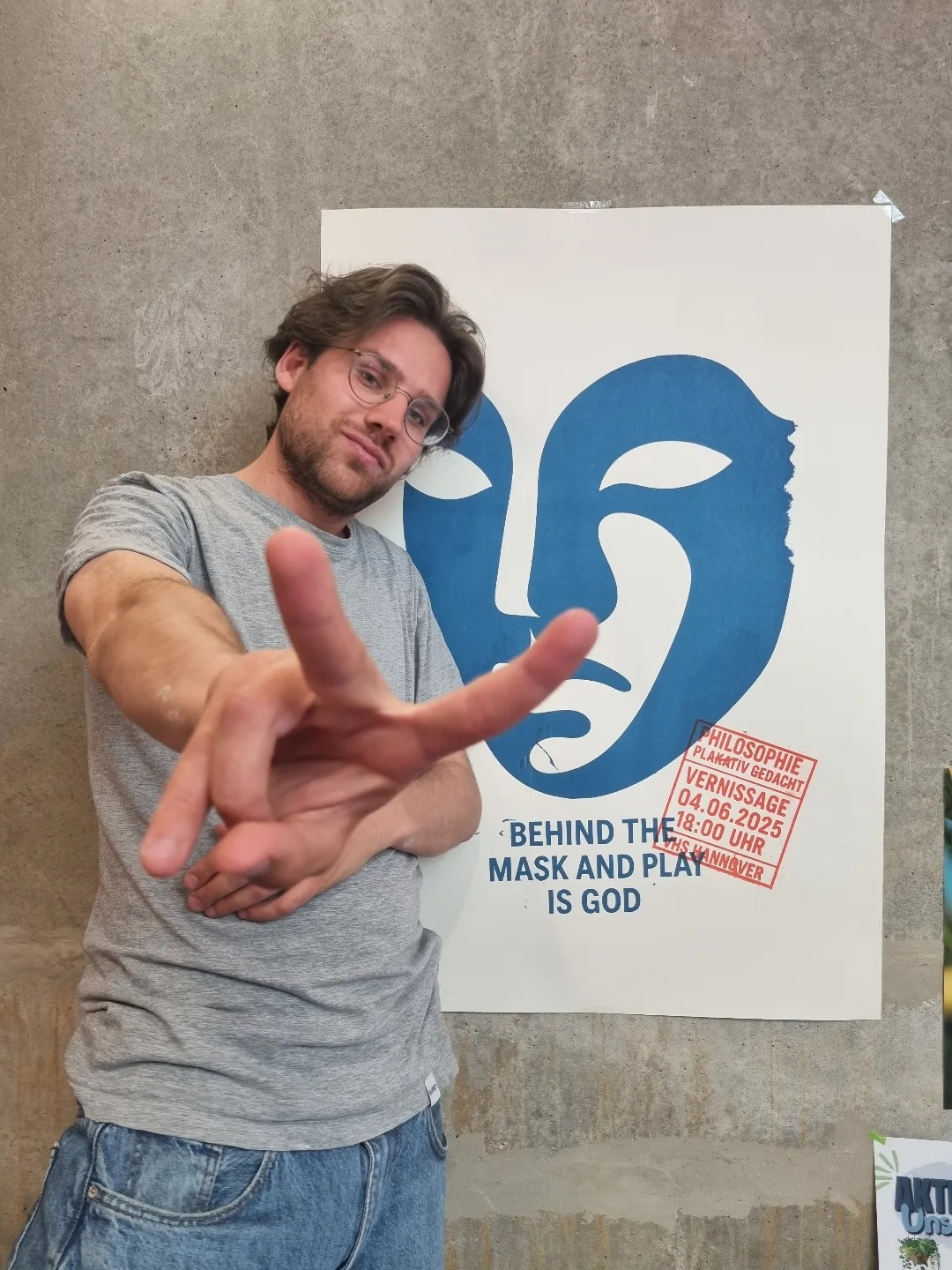 Lukas Wollny zeigt ein Peace-Zeichen vor seinem blauen Siebdruck Behind the Mask and Play is God